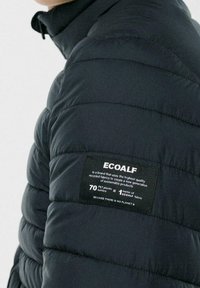 Černá prošívaná bunda s vysokým límcem a prošívanou texturou. Na levé straně u ramene je štítek s informacemi o recyklaci.