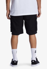 Schwarze Cargo-Shorts mit Seitentaschen, kombiniert mit weißen Socken und schwarzen Schuhen mit einem weißen Logo. Rückansicht des Outfits.