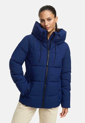Femme portant une veste rembourrée bleue avec un col haut, une fermeture éclair à l'avant et des poches à boutons sur les côtés, se tenant avec une main dans la poche.