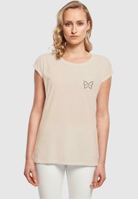 Beige bomulds T-shirt med korte ærmer, rund halsudskæring og et sort sommerfuglegrafik på venstre bryst. Glat tekstur, afslappet pasform.
