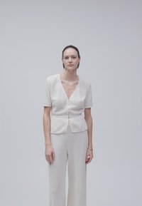 Γυναίκα με χτενισμένα προς τα πίσω μαλλιά, που φοράει λευκό υφασμάτινο κοντομάνικο τοπ με κουμπιά και ταιριαστό παντελόνι με φαρδιά μπατζάκια, που στέκεται μπροστά σε ένα απλό γκρι φόντο.