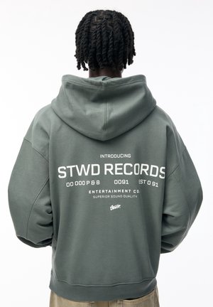 Osoba o krótkich skręconych włosach, ubrana w szarą bluzę z kapturem z napisem "STWD RECORDS" i powiązanym tekstem nadrukowanym na plecach.