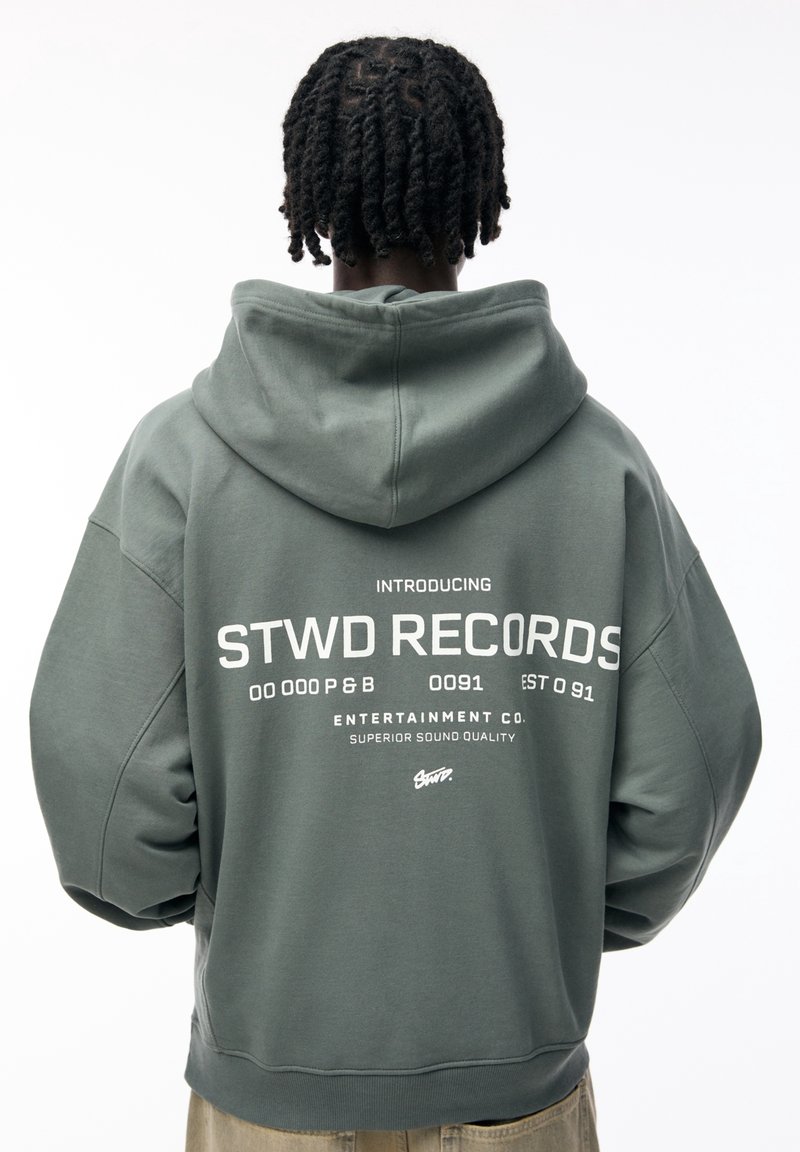 Personne aux cheveux courts torsadés portant un sweat à capuche gris avec « STWD RECORDS » et un texte associé imprimé dans le dos.