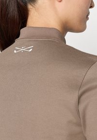 Brūns golfa polo krekls ar apkakli un teksturētu audumu. Augšējā aizmugurē ir redzams baltais Nike logo ar krustotiem golfa nūjām.