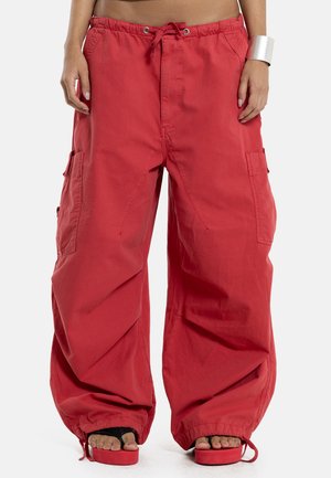 Wijde losse rode cargobroeken met aantrekkoord in de taille en elastische boorden gedragen door een staand persoon, gecombineerd met rode platformsandalen en een zilveren armband.
