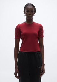 Piros, testhez simuló, rövid ujjú felső, rugalmas anyagból, kerek nyakkivágással és a gallér közelében diszkrét Calvin Klein logóval. Feket nadrág is látható.