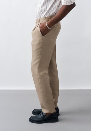 Uomo che indossa pantaloni beige su misura con pieghe, una camicia a righe, mocassini neri e un braccialetto d'argento, in piedi con una mano in tasca.