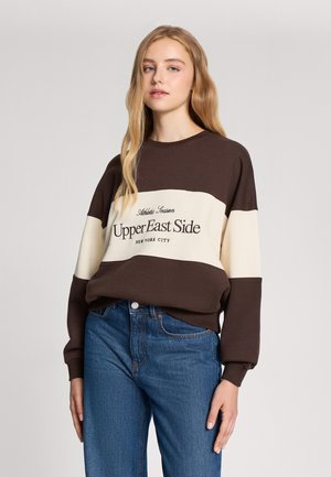 Felpa marrone e crema con scollatura rotonda, con testo "Upper East Side" in nero, abbinata a jeans blu a vita alta.