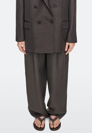 Personne portant un pantalon de costume marron foncé ample et un blazer croisé assorti, associé à des sandales marron sur fond clair.