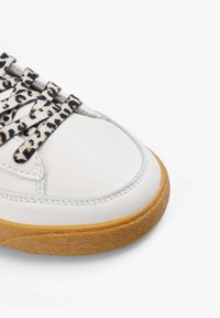 Zapatilla de cuero blanca con puntera redonda y suela de goma texturizada; cuenta con cordones de estampado de leopardo y costuras en la parte superior.