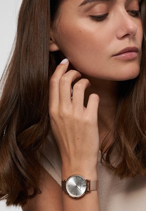 Nahaufnahme einer Frau mit langen braunen Haaren, die eine silberne Uhr mit einem Mesh-Armband am Handgelenk trägt und nachdenklich ihren Hals berührt.