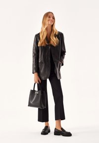 Schwarze Lederblazer mit einem schlanken, taillierten Design, kombiniert mit schwarze, cropped Hosen und klobigen Slippern. Das Modell hält eine schwarze Handtasche.