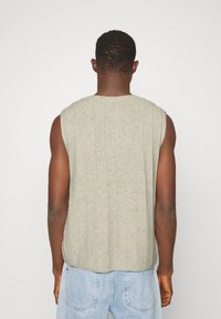 Maglione senza maniche grigio chiaro con una texture a coste, caratterizzato da un collo rotondo e sottili striature. Indossato sopra dei jeans blu chiaro.