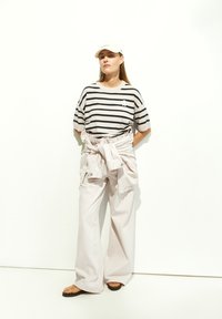 Gestreepte zwart-witte T-shirt, lichtroze wijde broek en een lichte pet. De outfit bevat een om de taille geknoopt jack.