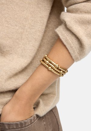 Zbliżenie na ramię z złotą bransoletką z koralików noszoną na beżowym swetrze z długim rękawem, ręka częściowo spoczywa w kieszeni brązowych spodni.