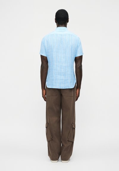 Man standing with back to camera wearing a light blue striped short-sleeve shirt and loose brown cargo pants with white shoes.