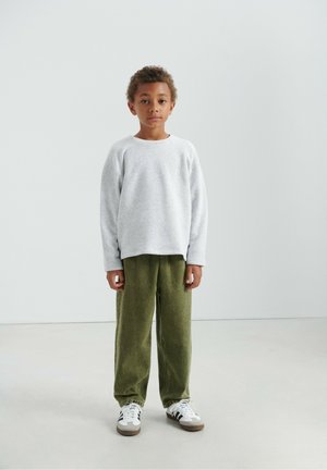 Sweat-shirt gris à manches longues et col ras du cou ; pantalon en velours côtelé vert ; baskets blanches avec des accents marron. Enfant debout devant un fond neutre.