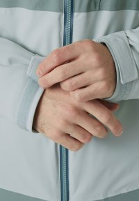 Lichtblauwe jas met een ritssluiting aan de voorkant, met een zachte textuur en contrasterende manchetten. Handen passen het manchetten-detail aan met een klittenbandstrap.