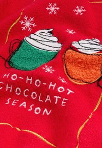 Tessuto rosso con tazze di cioccolata calda ricamate in verde e arancione, sormontate da panna montata bianca. Include dettagli di fiocchi di neve e testo.