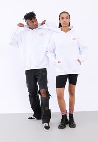 Felpe bianche oversize con tasche frontali, caratterizzate da un piccolo logo rosso. Jeans neri strappati e stivali neri fino a metà polpaccio abbinati a pantaloni biker neri.