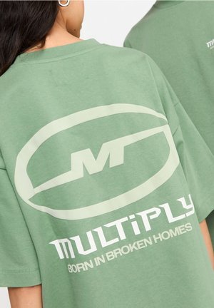 Woman wearing green oversized T-shirt with large white "Multiply" logo and text "Born in broken homes" on the back.