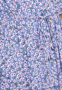 Ljusblå klänning med ett blommönster av vita och rosa prästkragar. Har en rynkad midja, dragsko och blå knappar. Bomullstyg.