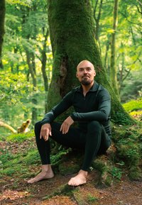 Czarna bluzka z długim rękawem na zamek i dopasowane legginsy. Siedzi boso oparta o porośnięte mchem drzewo w bujnym zielonym lesie.