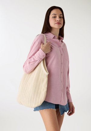 Camicia rosa chiaro con bottoni e tasca, abbinata a pantaloncini di jeans. L'individuo porta una borsa a rete color beige.