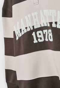 Smešani smeđi i svijetloružičasti prugasti dukserica s izvezenim natpisom "MANHATTAN 1978". Mekana tkanina s opuštenim krojem i detaljima šivanja.