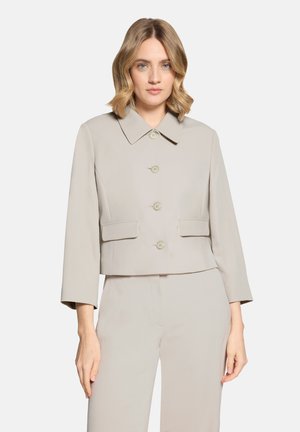 Vrouw met schouderlang blond haar, gekleed in een lichtbeige cropped jasje met knopen en bijpassende broek, tegen een witte achtergrond.