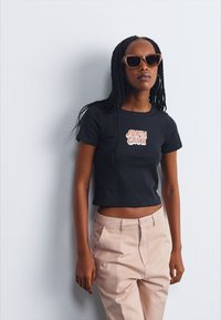 Svart kort t-skjorte med "SANTA CRUZ" grafikk i oransje, kombinert med lyse rosa bukser. Modellen har på seg solbriller og har flettet hår.