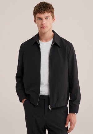 Ung mand iført en sort lynlåstrøje over en hvid t-shirt, med sorte bukser, stående mod en ensartet lys baggrund.