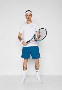 Camisa atlética branca com logotipo, calções teal e sapatos de desporto brancos. A segurar uma raquete de ténis. Os acessórios incluem uma faixa branca para a cabeça e faixas para os pulsos.