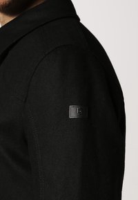 Cappotto di lana nera con un colletto strutturato, caratterizzato da un tessuto testurizzato e una toppa rettangolare con il marchio sulla manica sinistra.