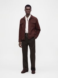 Homme portant une veste marron par-dessus une chemise blanche, un pantalon marron foncé et des chaussures marron, debout devant un fond clair uni.