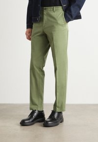 Uomo che indossa pantaloni verde oliva, scarpe nere stringate e una giacca di jeans scura, con una mano in tasca, in piedi su un pavimento neutro.