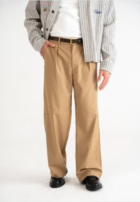 Beige, wijde broek gemaakt van een gladde stof met een lichte glans. Gecombineerd met een gestreepte jas, een bruine riem en zwarte schoenen.