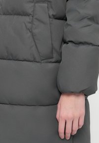 Hånden hviler ved lommen på en quilted mørkegrå polstret vinterjakke med syede horisontale sektioner.