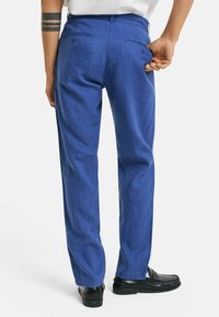Blauwe linnen broek met een rechte pasvorm, voorzien van twee achterzakken en een gladde textuur. Zwarte schoenen completeren de outfit.