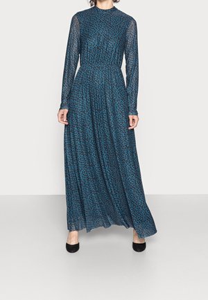 Femme portant une robe à manches longues, longueur cheville, avec un motif floral bleu et noir, associée à des chaussures noires, debout sur un sol clair.