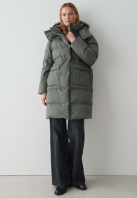 Parka vert olive bouffante avec capuche, poches avant et taille cintrée. Le matériau semble lisse avec une texture matelassée.