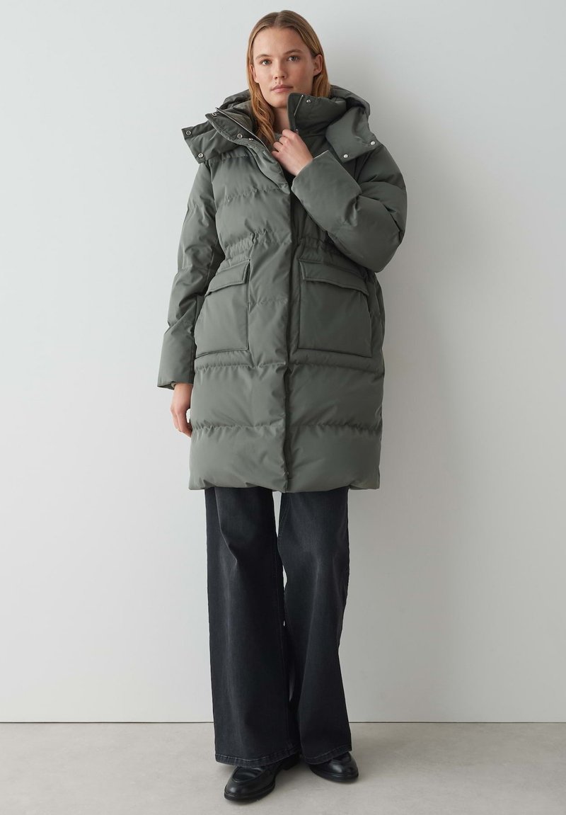 Parka vert olive bouffante avec capuche, poches avant et taille cintrée. Le matériau semble lisse avec une texture matelassée.