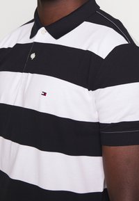 Polo rayé noir et blanc avec un col replié, présentant un petit logo sur la poitrine et des accents de couture contrastants.