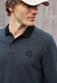 Dunkel gemustertes Poloshirt mit schwarzem Kragen. Verfügt über eine strukturierte Webart und ein quadratisches Logo-Patch auf der linken Seite.