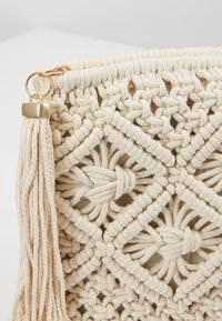 Motif tissé en macramé de couleur crème avec un détail de pompon et d'anneau doré sur une surface en tissu.