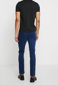 Man med svart kortärmad skjorta, blå slim-fit byxor och svarta kostymskor, stående vänd bort från kameran framför en neutral vit bakgrund.