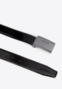 Zwarte leren riem met een zilveren metalen gesp. De gesp heeft een gestructureerd oppervlak en de gegraveerde merknaam "WITTCHEN."