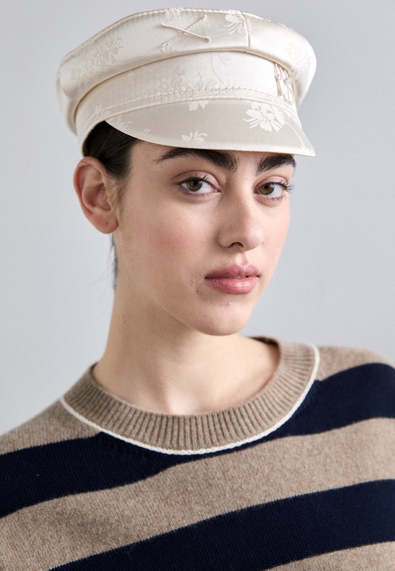 Off-white fabric cap features floral embroidery and a structured visor. Model wears a brown and navy striped sweater with a ribbed neckline.