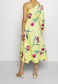 Žena v žlutých midi šatech na jedno rameno s růžovým potiskem třešňových květů a jedním dlouhým rukávem, spárovanými s béžovými sandály na blokovém podpatku.