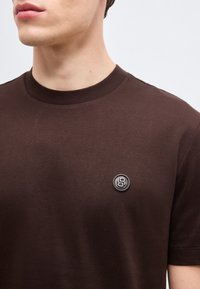 Άνδρας που φοράει καφέ σκούρο μπλουζάκι με στρογγυλή μαύρη έμβλημα με τον αριθμό 8 στο αριστερό στήθος.